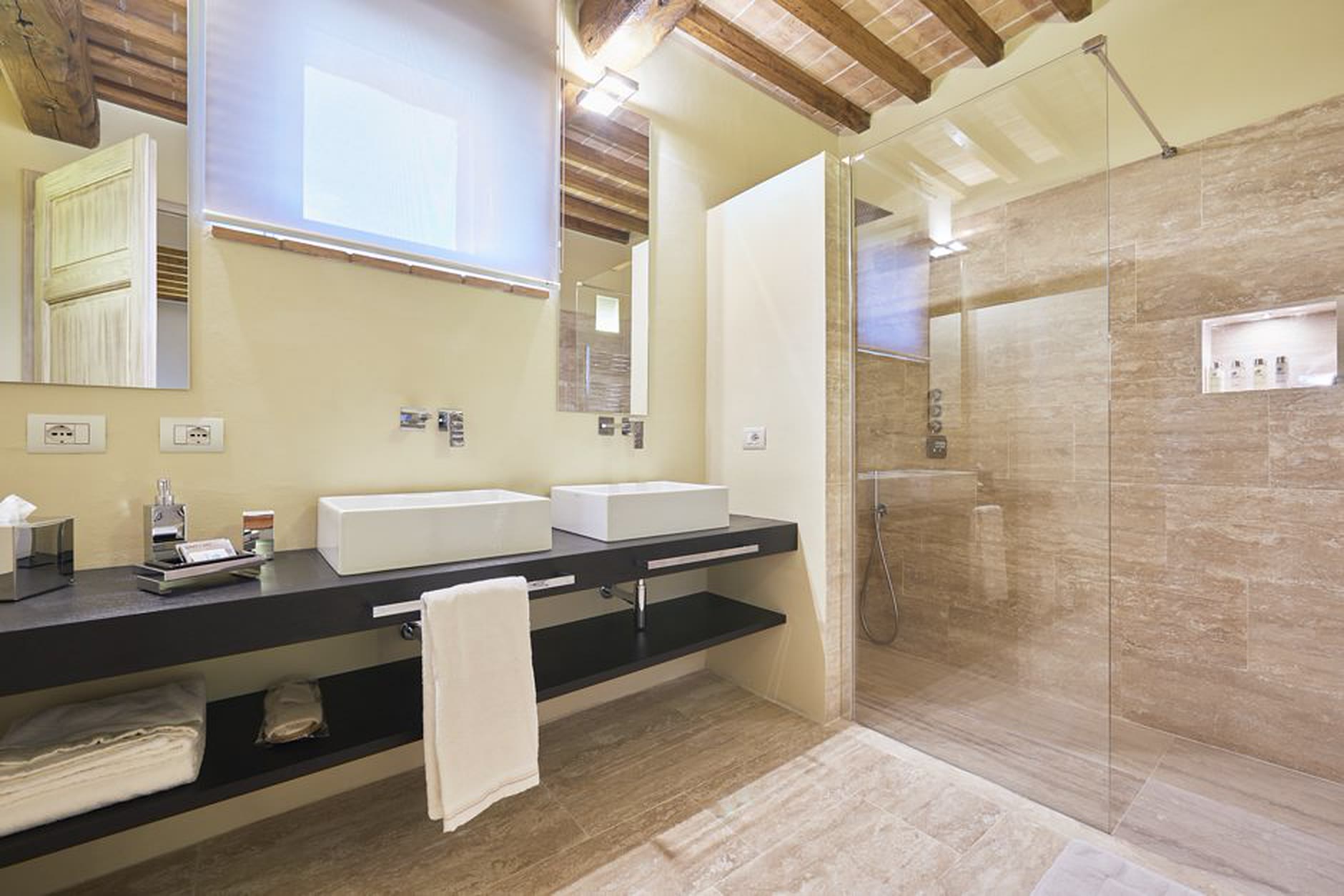 Bagno in travertino con doppio lavabo su mensola nera, doccia walk-in e soffitto in legno: materia calda e luce naturale.