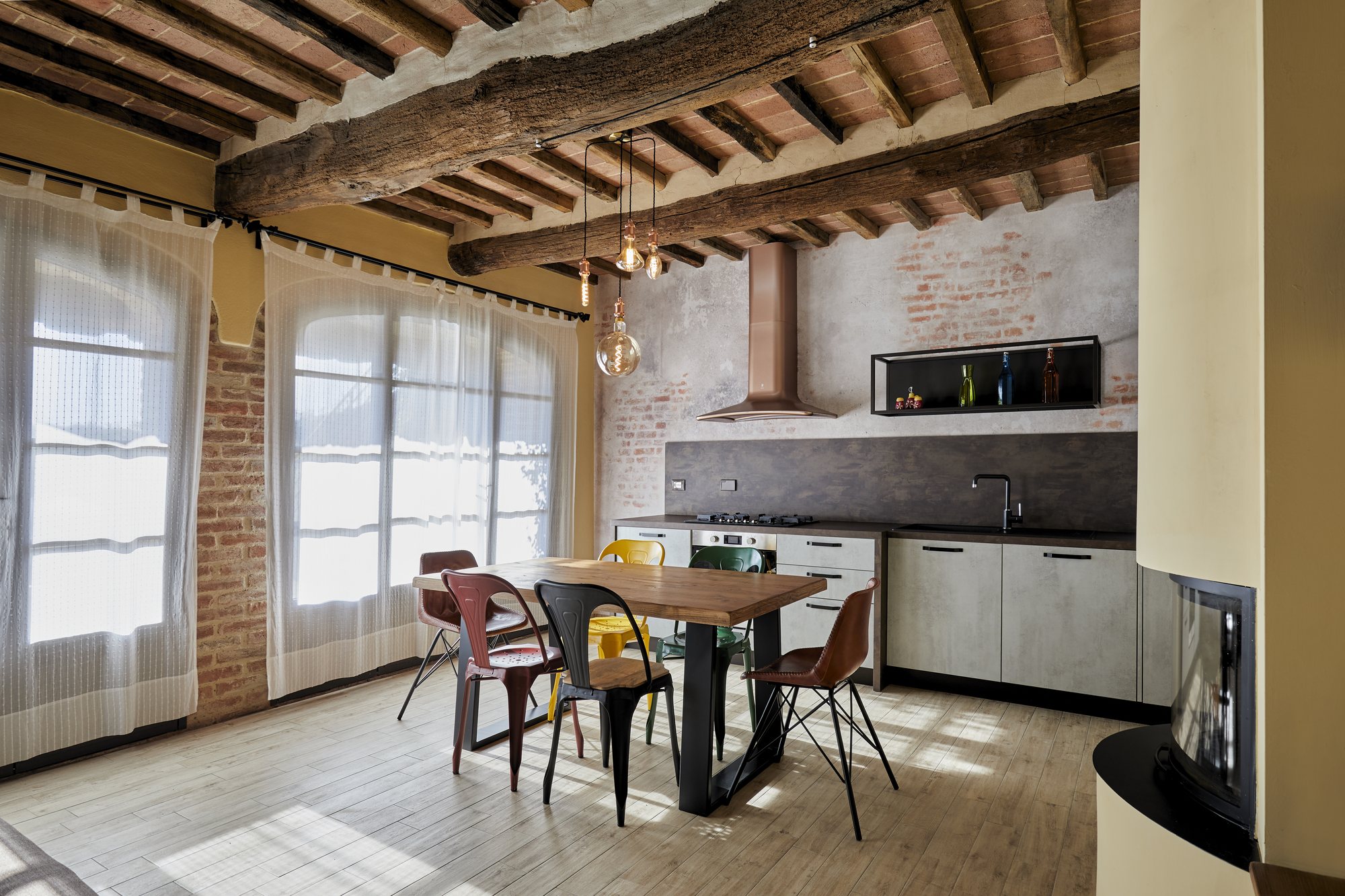Cucina in un casale ristrutturato: basi effetto cemento sotto travi antiche, cappa color rame, mattoni a vista e tavolo in legno con sedie in metallo colorato.