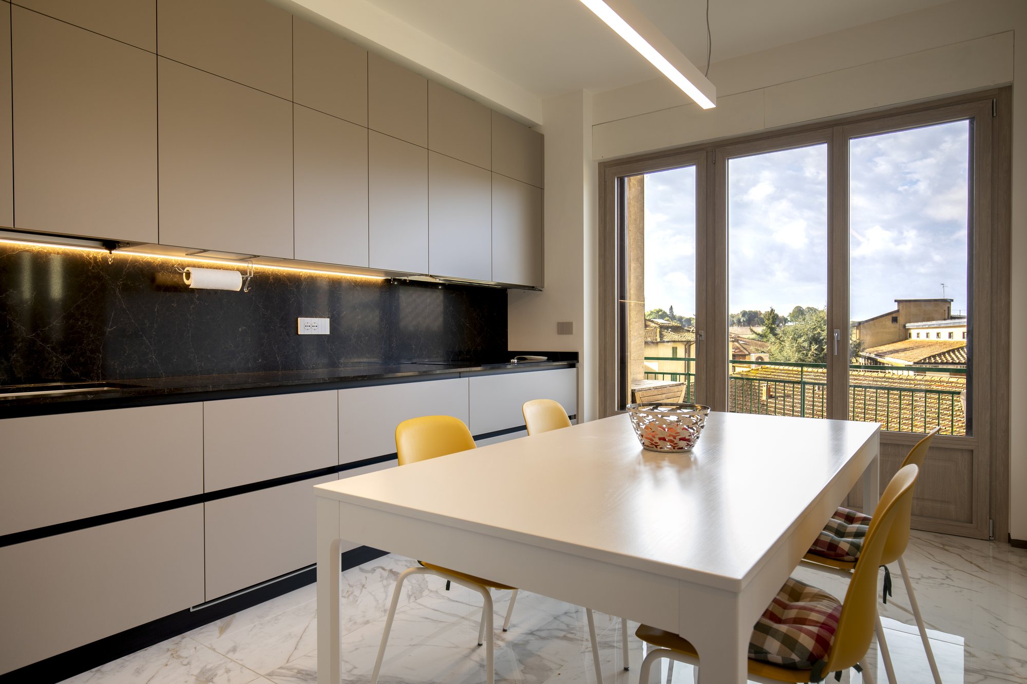 Cucina tortora con schienale nero retroilluminato, tavolo bianco e sedie gialle davanti alla portafinestra sui tetti.