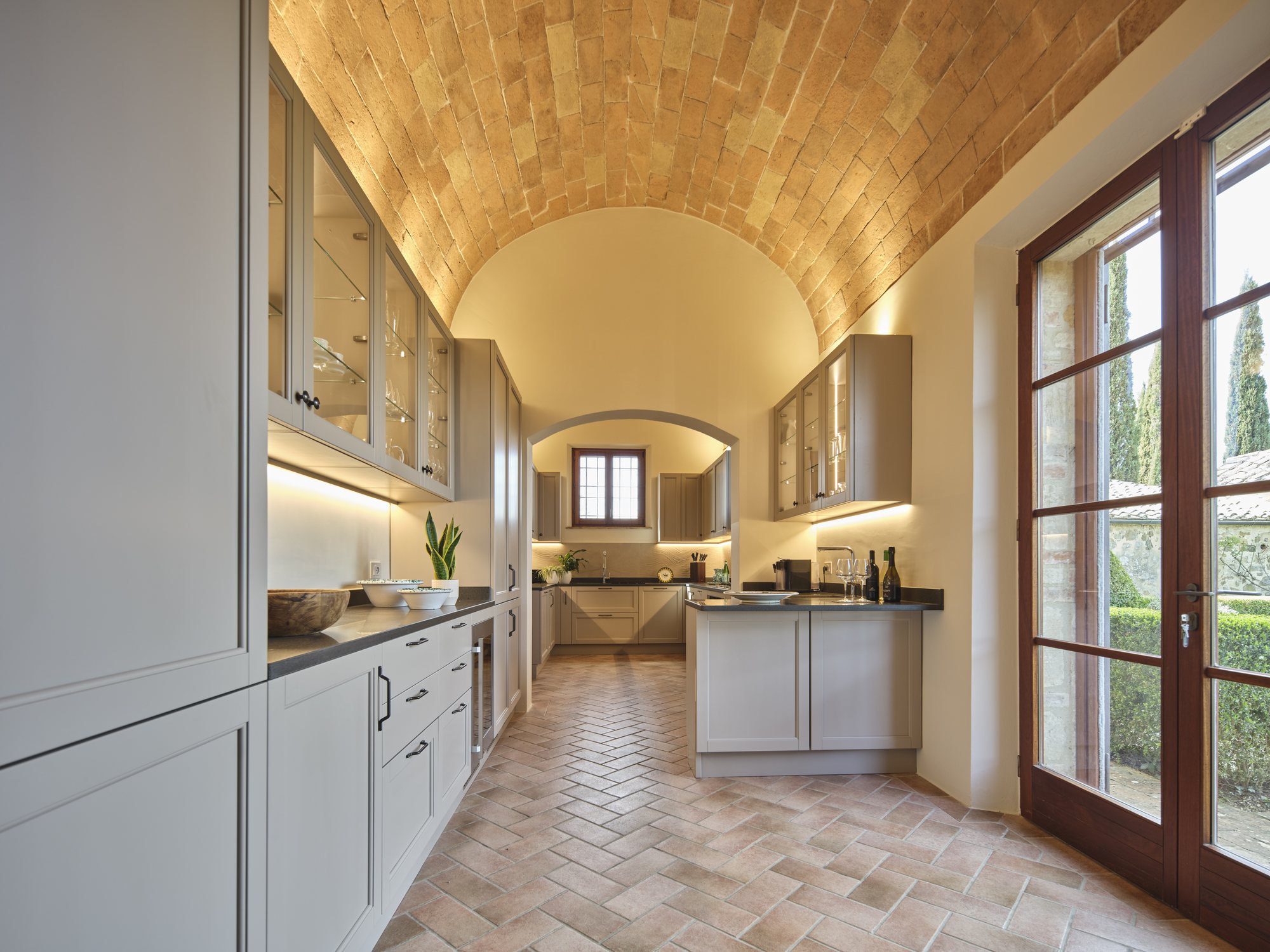 Cucina stile shaker grigio tortora sotto una volta a botte in cotto originale, pavimento a spina di pesce e portafinestra sul giardino.