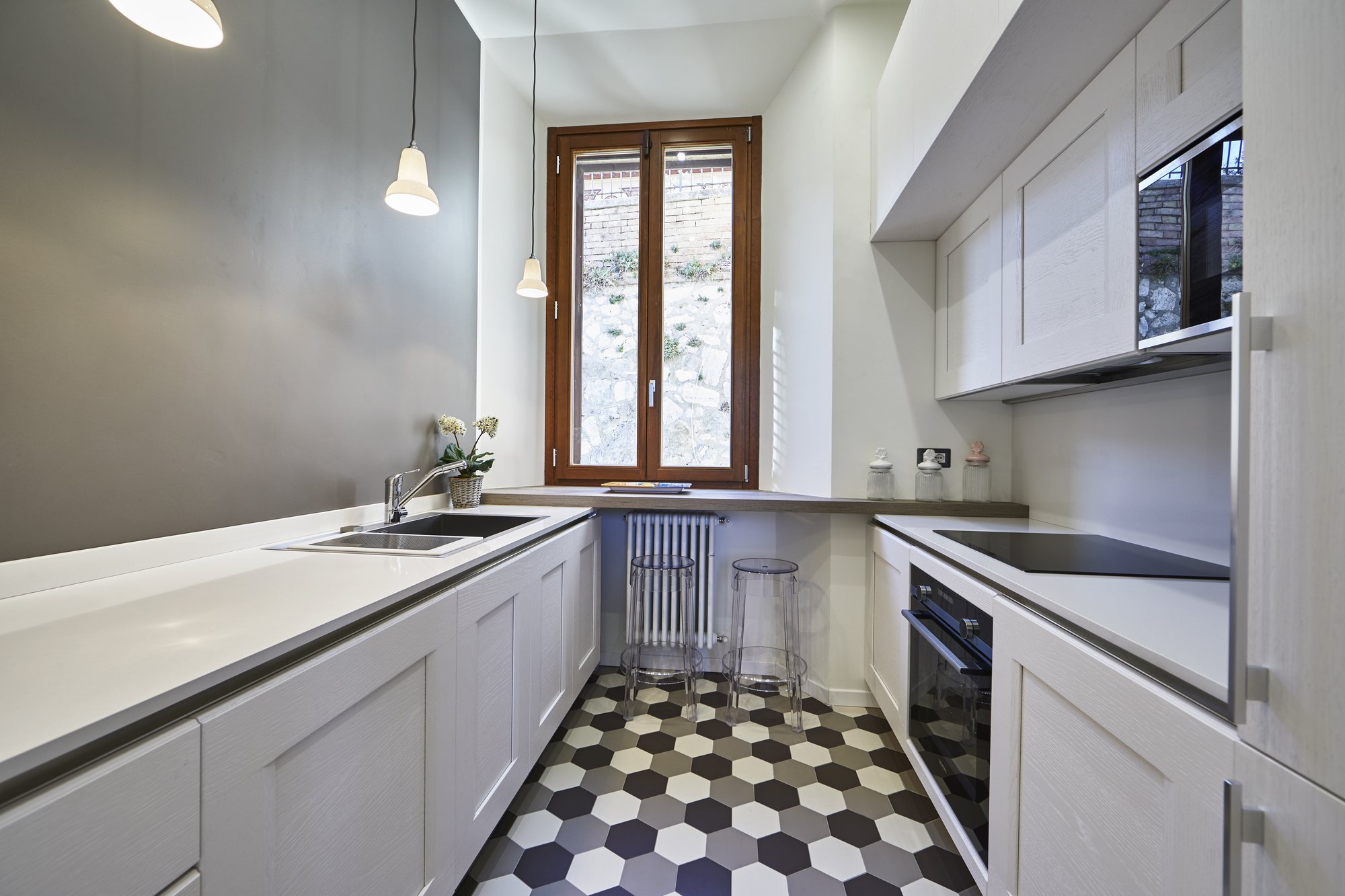Cucina a parallelo bianca con piano continuo, pavimento a esagoni e finestra in legno sul verde: il ritmo della geometria.