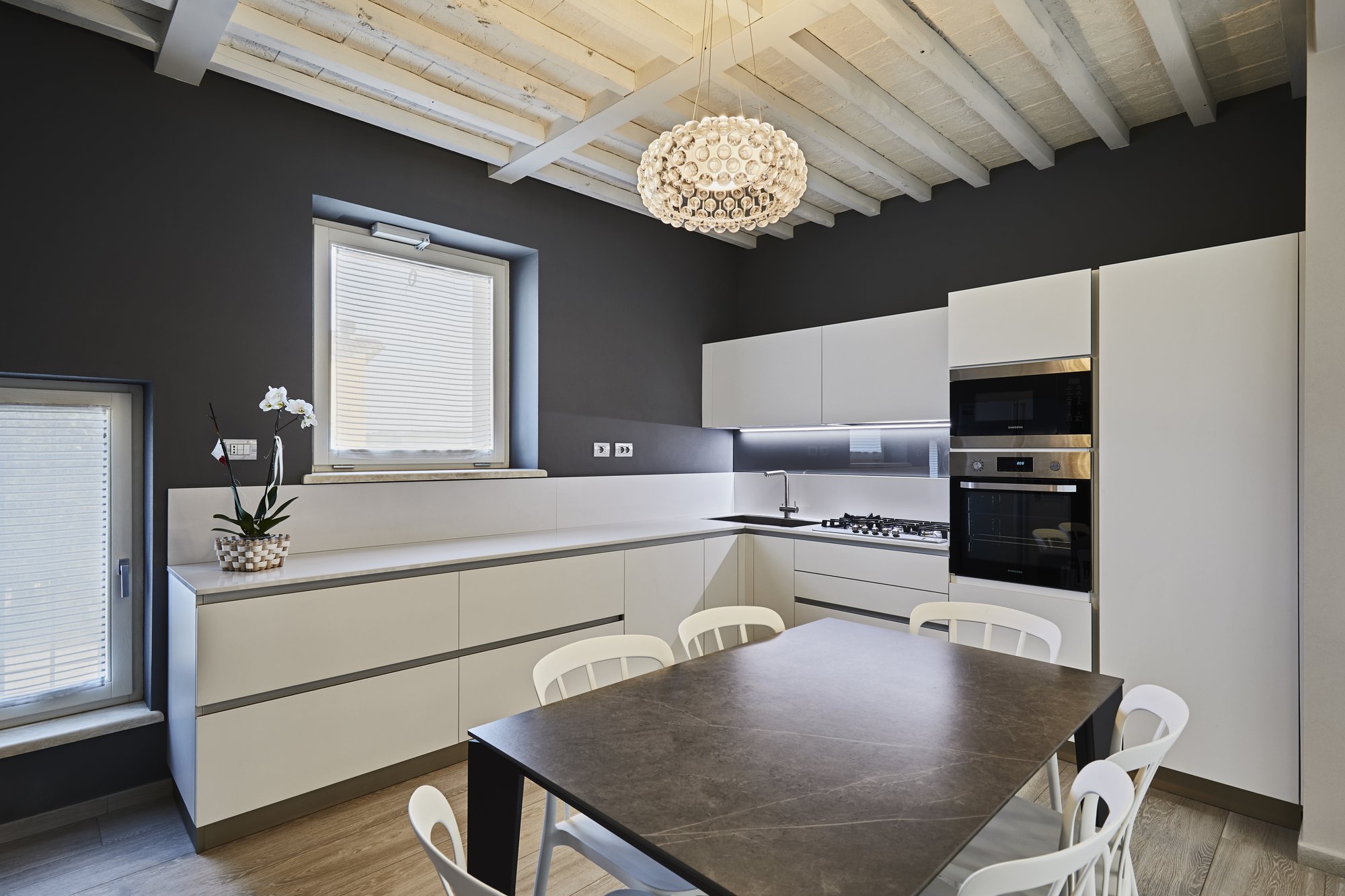 Cucina bianca opaca su parete antracite, travi sbiancate, lampadario scenografico e tavolo in gres: contrasto pulito e luce naturale.