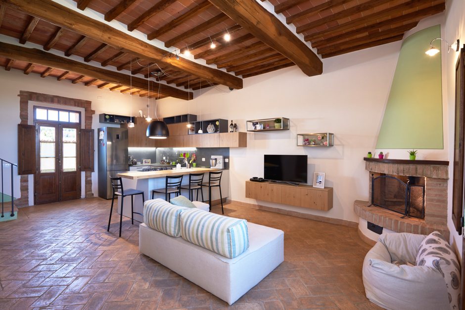 Zona giorno open space in casale: cucina con isola e sgabelli, camino d'angolo in mattoni, divano chiaro su pavimento in cotto.