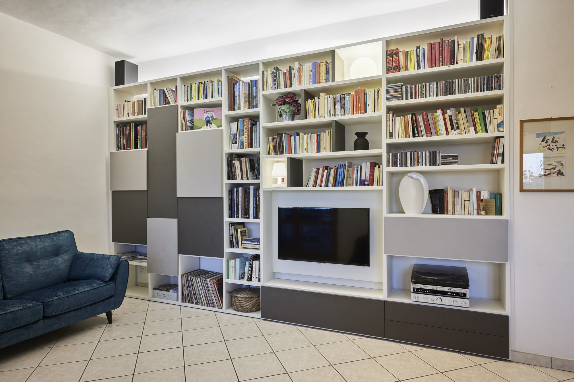 Parete attrezzata bianca e grigia su misura con vano TV, divano blu e libreria integrata.