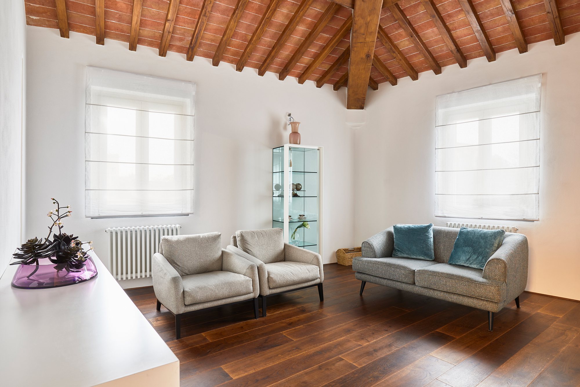 Salotto elegante sotto tetto in travi e mezzane: coppia di poltrone e divano grigio, vetrina di design e parquet scuro.