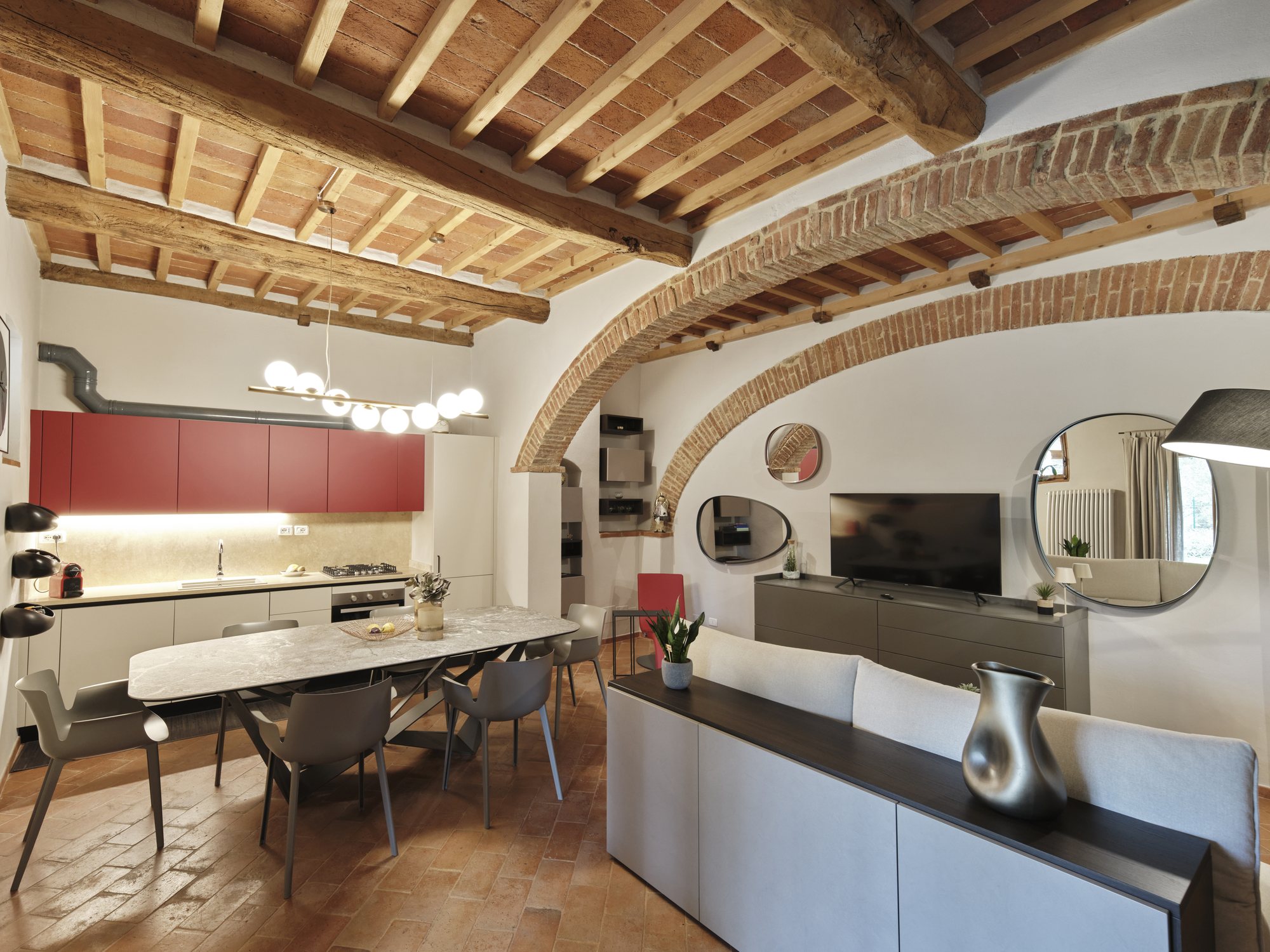 Open space con travi e archi in mattoni originali, cucina laccata rossa, tavolo in marmo e zona living: antico e contemporaneo insieme.