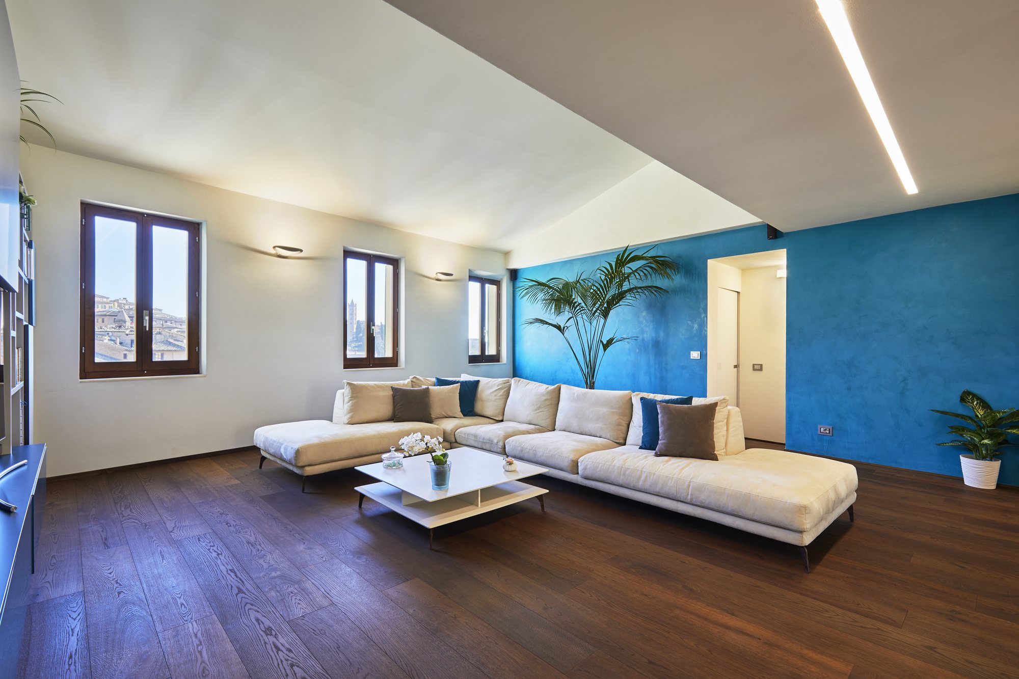 Ampio soggiorno con divano angolare champagne, parete blu intenso spatolato, pianta tropicale e parquet scuro.