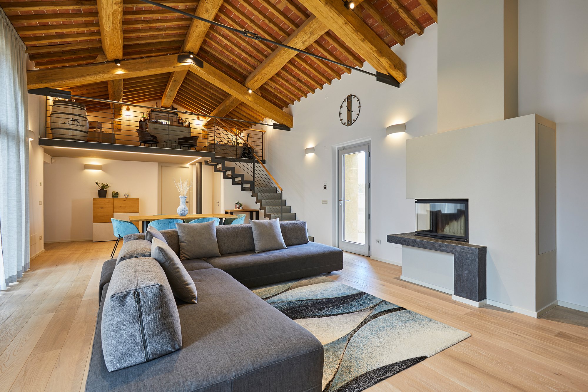 Grande living a doppia altezza con capriate in legno, soppalco, divano angolare grigio e camino moderno.