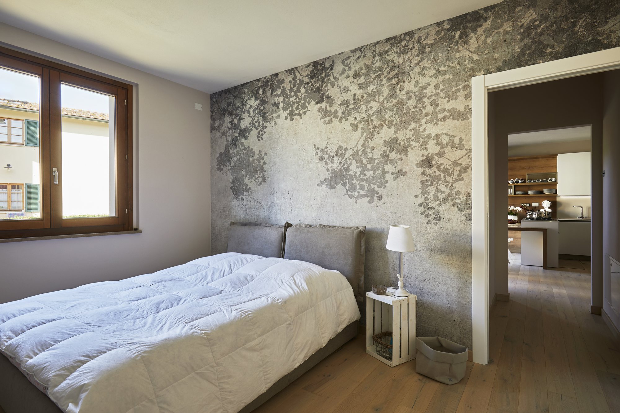 Camera matrimoniale con carta da parati a fogliame grigio, letto imbottito e comodino in cassetta di legno bianca: naturale e sereno.