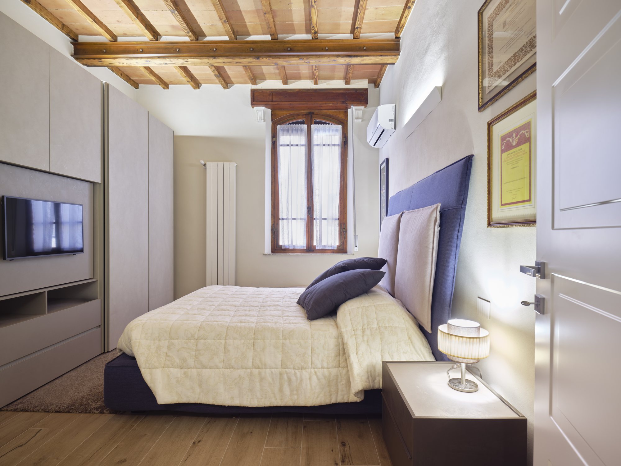 Camera in palazzo storico: testiera imbottita, parete attrezzata con TV e soffitto in travi e mezzane restaurato.