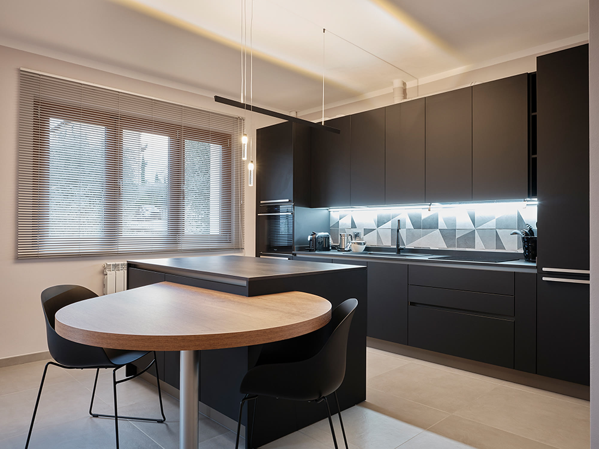 Cucina nera opaca con tavolo tondo in legno e luce lineare sulla parete attrezzata.