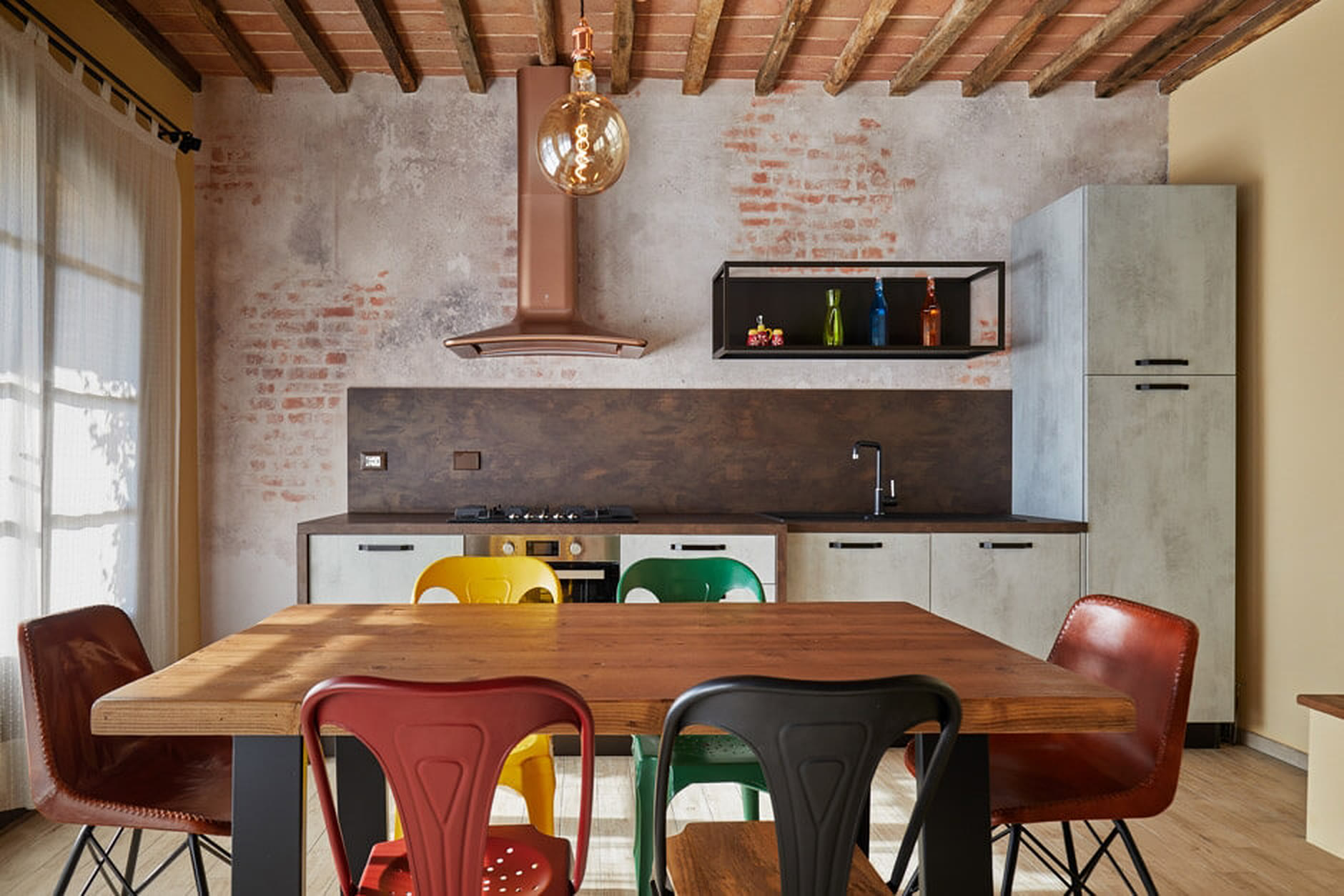 Cucina industrial con parete in mattoni, cappa ramata e sedie in metallo colorato.