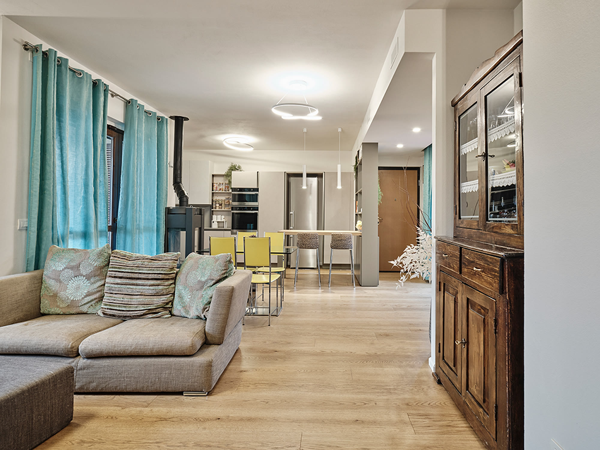 Open space con divano beige, stufa e cristalliera d'epoca: moderno e memoria insieme.