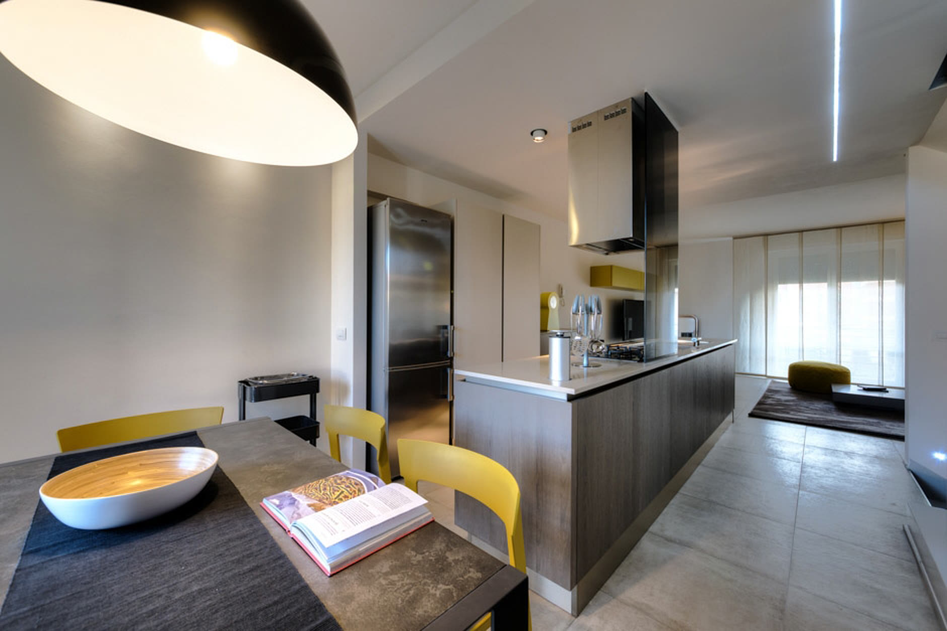 Cucina scura con isola, sedie gialle e grande lampada a campana.